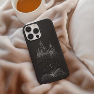 May include: Black phone case featuring a white line drawing of a castle, an open book, and stars. The case is lying on a soft, beige blanket next to a white mug filled with a brown liquid.