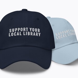 Peut inclure: Deux casquettes de baseball, une bleu marine et une bleu clair, sont présentées. Les deux casquettes portent l'inscription brodée en blanc "SUPPORT YOUR LOCAL LIBRARY". Les casquettes ont un design classique à six panneaux.