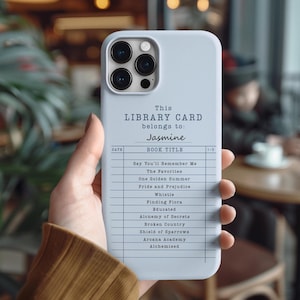 May include: A light blue phone case featuring a library card design. The text reads "This Library Card belongs to: Jasmine" with a list of book titles below. The phone case is held in a person's hand.