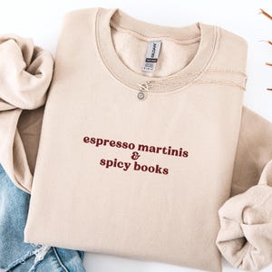 Pode incluir: Camisola bege com gola redonda com o texto "espresso martinis & spicy books" em fonte vermelho escuro. Um colar de prata com um pingente de sol está na camisola. A camisola está dobrada sobre uma superfície branca.