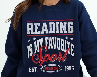 May include: Navy blue sweatshirt with the text "READING IS MY FAVORITE SPORT" in white and red lettering. The design includes stars and the words "EST. READ ON 1995". A comfortable and stylish garment for book lovers.