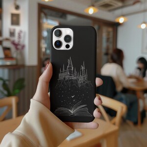 May include: Black phone case featuring a white line drawing of a castle on a hill, with an open book at the bottom. The case has a glossy finish and is designed for a smartphone. The case is held in a person's hand.