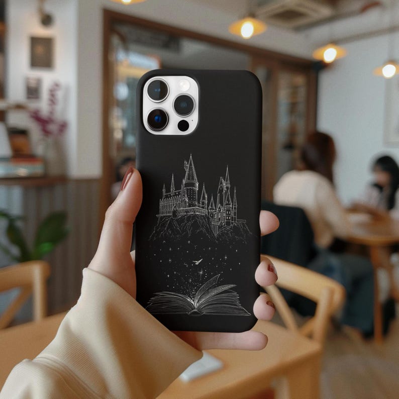 May include: Black phone case featuring a white line drawing of a castle on a hill, with an open book at the bottom. The case has a glossy finish and is designed for a smartphone. The case is held in a person's hand.