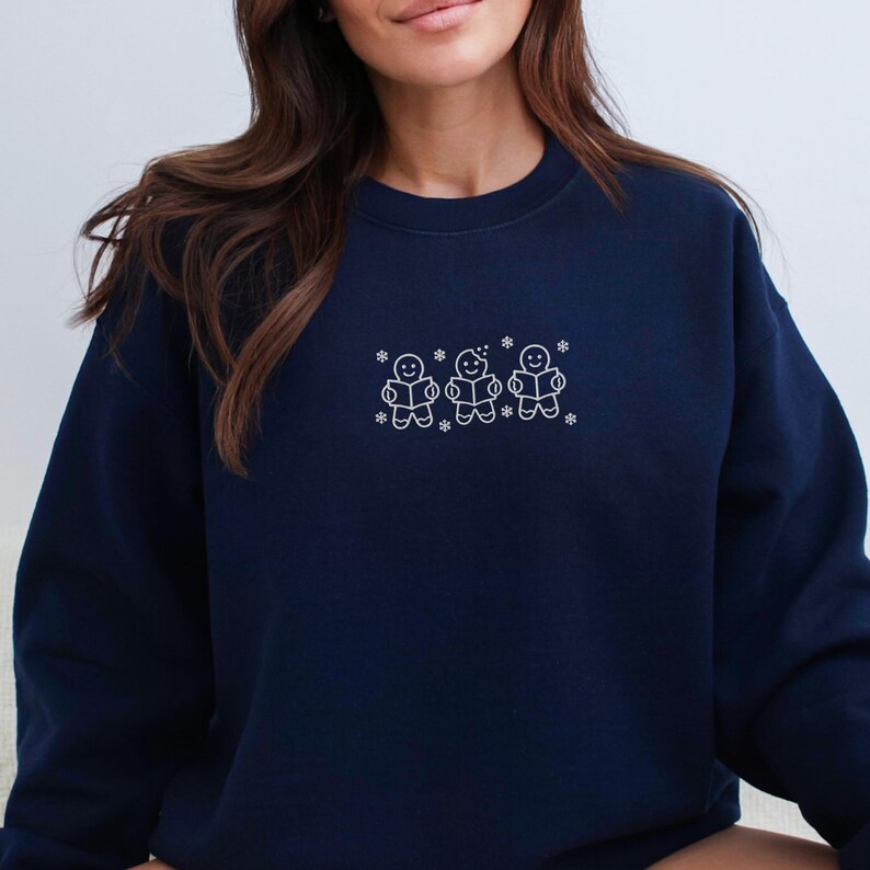 Pode incluir: Camisola azul marinho com um desenho bordado branco de tr&ecirc;s bonecos de gengibre lendo livros, com flocos de neve. Gola redonda.