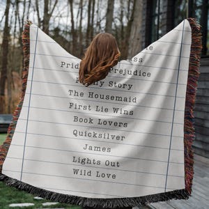 May include: A cream-colored throw blanket with a lined paper design and a list of book titles printed in black. The blanket has a fringed edge with a mix of colors. Titles include "Pride & Prejudice" and "First Lie Wins."