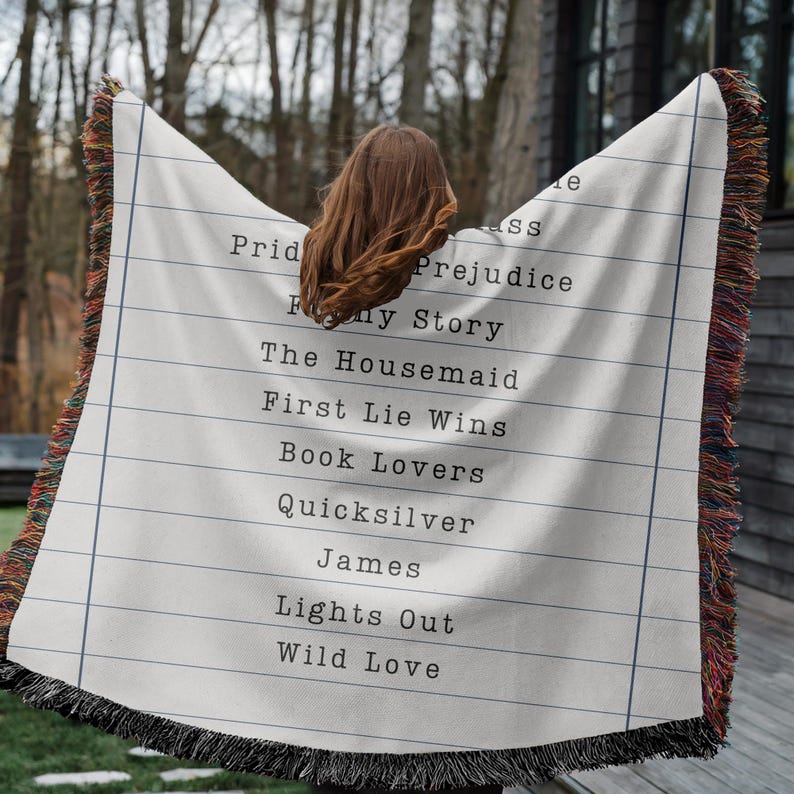 May include: A cream-colored throw blanket with a lined paper design and a list of book titles printed in black. The blanket has a fringed edge with a mix of colors. Titles include "Pride & Prejudice" and "First Lie Wins."