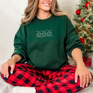 Pode incluir: Uma camisola verde escuro com um desenho bordado de tr&ecirc;s bonecos de gengibre. A pessoa est&aacute; a usar cal&ccedil;as de pijama xadrez vermelhas e pretas. Uma &aacute;rvore de Natal &eacute; vis&iacute;vel no fundo.