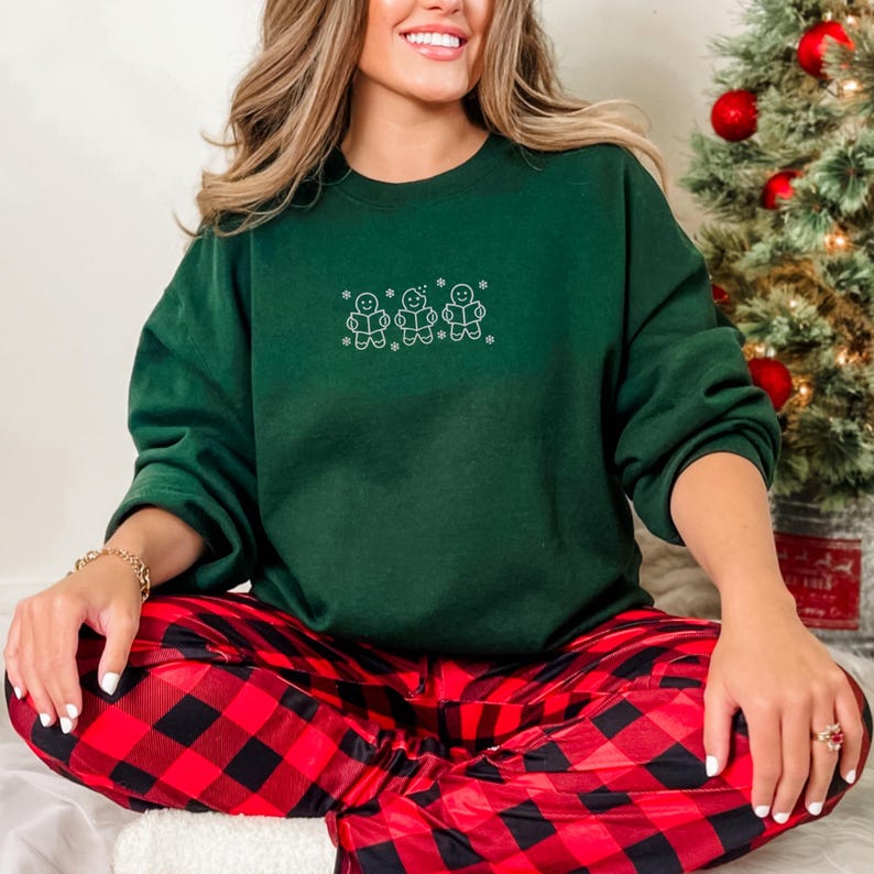 Pode incluir: Uma camisola verde escuro com um desenho bordado de tr&ecirc;s bonecos de gengibre. A pessoa est&aacute; a usar cal&ccedil;as de pijama xadrez vermelhas e pretas. Uma &aacute;rvore de Natal &eacute; vis&iacute;vel no fundo.