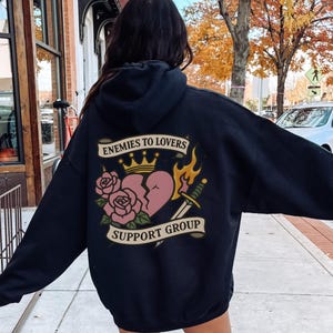 Pode incluir: Um moletom preto com um gráfico nas costas. O gráfico inclui um coração partido, rosas, uma coroa, uma adaga e as palavras "ENEMIES TO LOVERS" e "SUPPORT GROUP" em uma faixa.