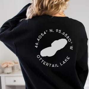 May include: Black sweatshirt with a white graphic of Ottertail Lake, including coordinates 46.4054&deg; N, 95.6580&deg; W. The lake outline and text are in a circular design. The sweatshirt is a casual style.