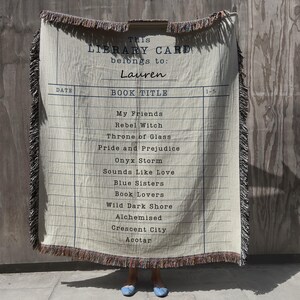 May include: Cream-colored throw blanket with a library card design. The blanket features a list of book titles and the text "This Library Card belongs to: Lauren." The edges are fringed with brown and pink threads. A unique gift for book lovers.