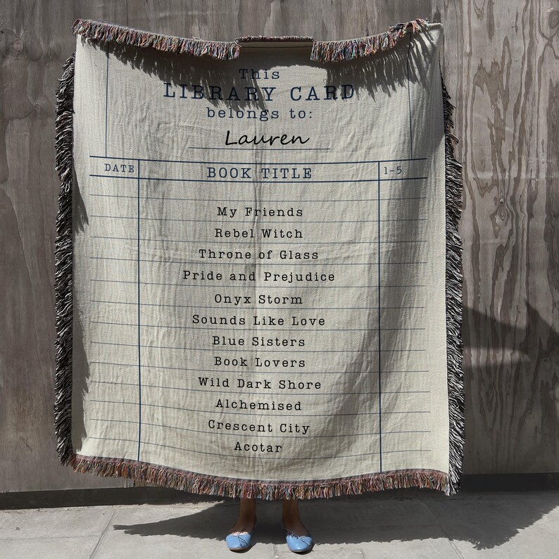 May include: Cream-colored throw blanket with a library card design. The blanket features a list of book titles and the text "This Library Card belongs to: Lauren." The edges are fringed with brown and pink threads. A unique gift for book lovers.