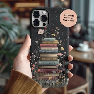 Op de afbeelding: Zwarte telefoonhoes met een ontwerp van opgestapelde boeken, waaronder titels als "Pride" en "Little Women". De hoes is versierd met bloemen in roze, geel en wit. Tekst op de hoes luidt "Customize for your fave titles!"
