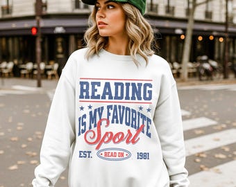 May include: White sweatshirt with the text "READING IS MY FAVORITE SPORT" in blue and red. The sweatshirt also has the text "EST. READ ON 1981". The person in the image is wearing a green baseball cap.