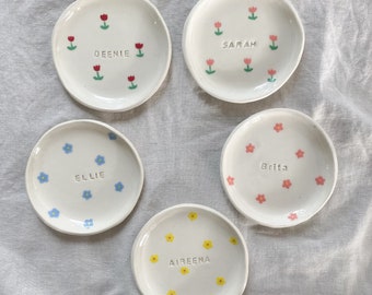 May include: Five small white ceramic dishes with different floral designs and names written on them. The names are Deenie, Sarah, Ellie, Britta, and Aireena.