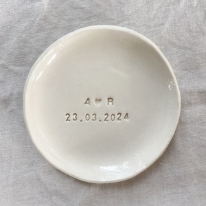 May include: A round, off-white ceramic trinket dish with a smooth, glossy finish. The centre of the dish is stamped with the letters A and R separated by a small heart symbol, followed by the date 23.03.2024 embossed in the clay surface.