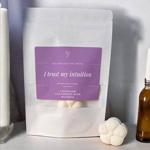 May include: A white pouch with a purple label that reads "I trust my intuition" and "Restore Your Wellbeing". The pouch contains shower steamers. A white, flower-shaped shower steamer sits next to the pouch.