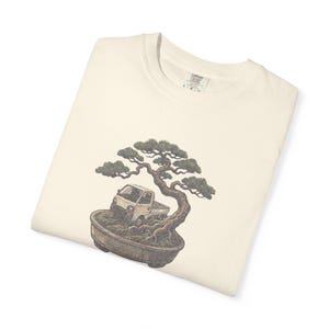 May include: An off-white t-shirt featuring a graphic print of a small white pickup truck parked inside a ceramic bonsai pot beneath a stylized green and brown tree. The shirt has a crew neckline and a visible interior label at the collar.