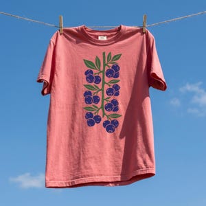 May include: A coral-coloured t-shirt with a green and blue blueberry design hanging on a clothesline against a blue sky. The graphic features a vertical arrangement of blueberries and leaves.
