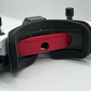 Könnte beinhalten: Eine weiße und schwarze FPV-Brille mit einer roten "Vor dem Flug entfernen"-Abdeckung über dem Objektiv.