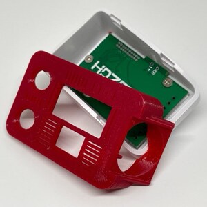 May include: A red 3D printed case with a cutout for a circuit board. The case has a rectangular shape with rounded corners and several holes. The circuit board is green and has the text "HDZ" printed on it.