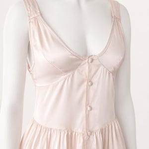 May include: A pale pink sleeveless dress with a deep scoop neckline and a button-down front featuring matching fabric-covered buttons. The bodice has structured seams, and the skirt portion includes a horizontal tiered ruffle detail.