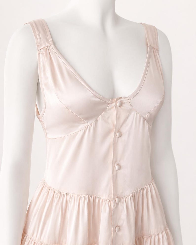 May include: A pale pink sleeveless dress with a deep scoop neckline and a button-down front featuring matching fabric-covered buttons. The bodice has structured seams, and the skirt portion includes a horizontal tiered ruffle detail.