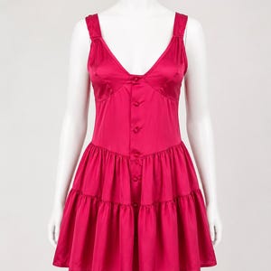 May include: A vibrant magenta satin mini dress featuring a deep V-neckline, wide shoulder straps, and a front button-down closure. The bodice is fitted, transitioning into a gathered, tiered skirt that creates a flared silhouette with a smooth finish.