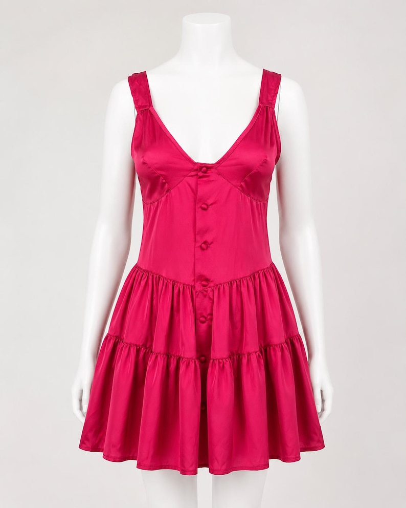 May include: A vibrant magenta satin mini dress featuring a deep V-neckline, wide shoulder straps, and a front button-down closure. The bodice is fitted, transitioning into a gathered, tiered skirt that creates a flared silhouette with a smooth finish.