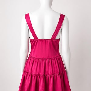 May include: A vibrant magenta sleeveless dress featuring a square neckline and wide shoulder straps. The garment is constructed with a fitted bodice that transitions into a two-tiered, gathered skirt, creating a flared silhouette with a smooth finish.