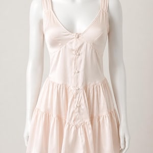May include: A pale pink sleeveless mini dress featuring a deep V-neckline, a button-down front with five matching fabric-covered buttons, and a tiered skirt design. The garment is crafted from a smooth, satin-like fabric with a subtle sheen.