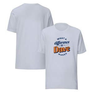 Può includere: Una t-shirt blu chiaro con una stampa grafica bianca che dice "What a difference a Dave makes". Il testo è in uno stile di font retrò con colori arancioni e blu.