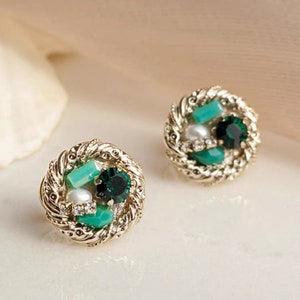 May include: A pair of gold-tone earrings with a circular design. The earrings feature a cluster of green and white stones set in a twisted rope-like frame.