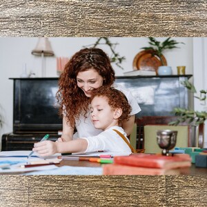 May include: A woman and a child are sitting at a table, coloring on a piece of paper. The woman has long, curly red hair and is wearing a white shirt. The child has short, curly brown hair and is wearing a white shirt. There are colored pencils and markers on the table.