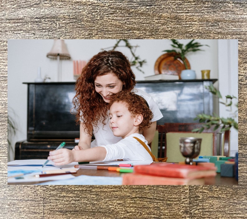 May include: A woman and a child are sitting at a table, coloring on a piece of paper. The woman has long, curly red hair and is wearing a white shirt. The child has short, curly brown hair and is wearing a white shirt. There are colored pencils and markers on the table.