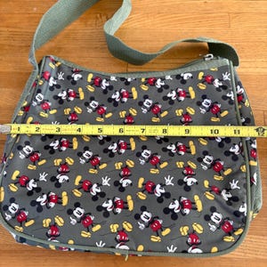 Puede incluir: Un bolso de hombro gris con un patr&oacute;n repetitivo de Mickey Mouse en rojo, negro y amarillo. Una cinta m&eacute;trica amarilla Stanley est&aacute; colocada sobre el frente, mostrando un ancho de aproximadamente 29,2 cm. Tiene una correa ajustable verde.