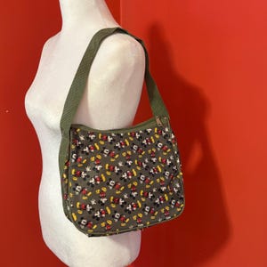 Puede incluir: Un bolso de hombro con base de color verde oliva y un patr&oacute;n repetitivo de figuras de Mickey Mouse en rojo, amarillo, negro y blanco. El bolso incluye una correa de hombro de tela verde oliva a juego y un cierre de cremallera superior.