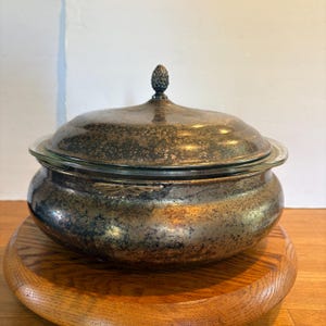 May include: A round silver-plated serving dish with a matching lid featuring a decorative pinecone-shaped finial. The metal surface displays a dark, aged patina with visible tarnishing throughout the exterior of the bowl and the domed lid.