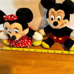 May include: Two plush Disney characters, Mickey and Minnie Mouse, sit side-by-side. Minnie wears a red polka-dot dress and bow. Mickey wears red shorts and yellow shoes. A yellow tape measure is in the foreground.