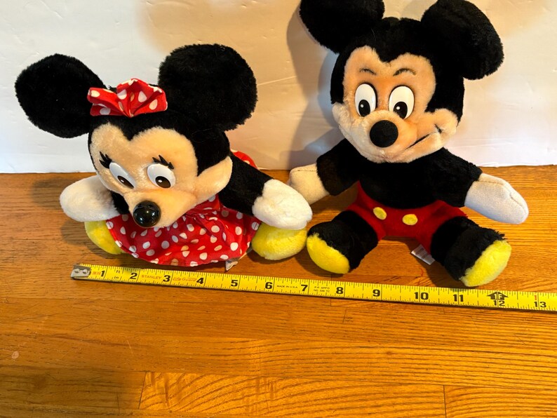 May include: Two plush Disney characters, Mickey and Minnie Mouse, sit side-by-side. Minnie wears a red polka-dot dress and bow. Mickey wears red shorts and yellow shoes. A yellow tape measure is in the foreground.