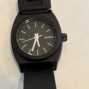 May include: Black Nixon watch with a black band. The watch face is black with white hands and markers. The word "NIXON" is printed in white on the watch face. The watch has a black crown.