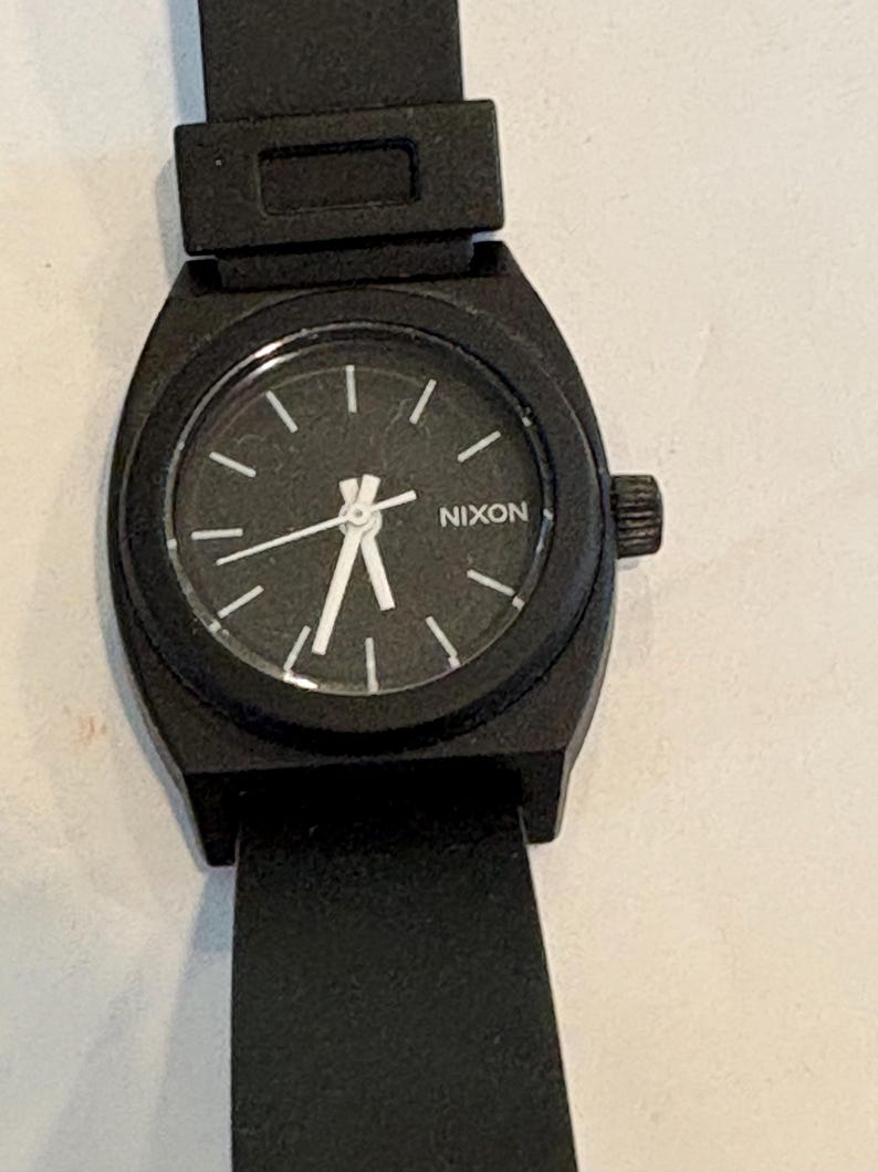 May include: Black Nixon watch with a black band. The watch face is black with white hands and markers. The word "NIXON" is printed in white on the watch face. The watch has a black crown.