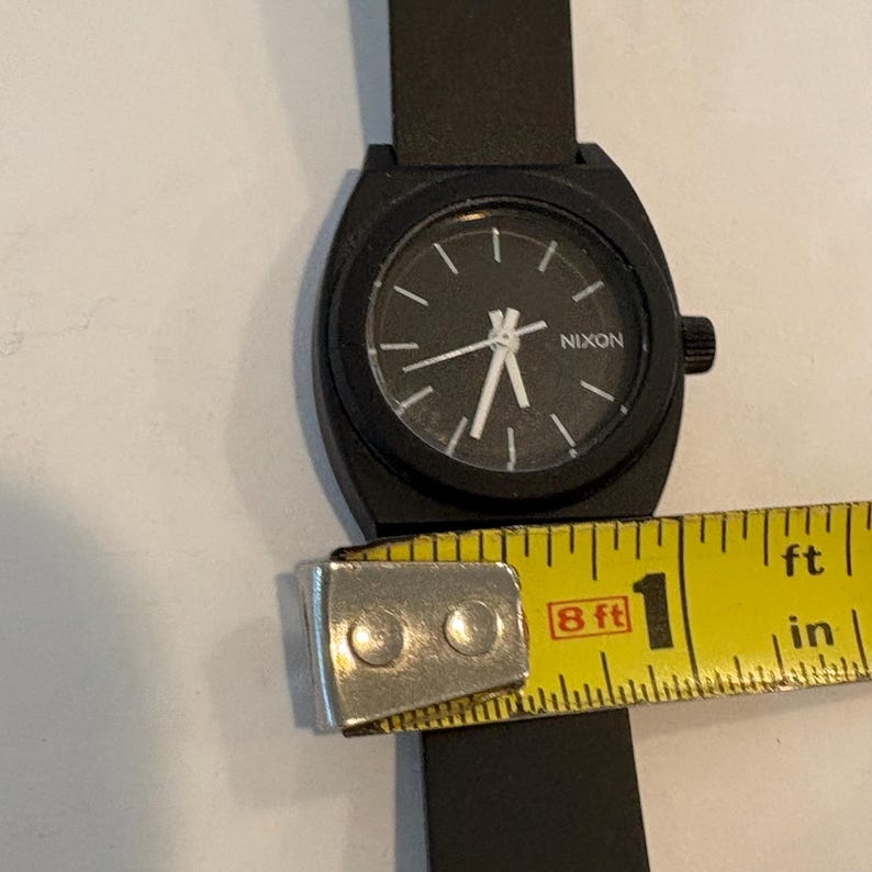 May include: A black Nixon watch with a black band. The watch face is black with white hands and markers. The watch is approximately 1 inch in diameter. The word "NIXON" is printed on the watch face.