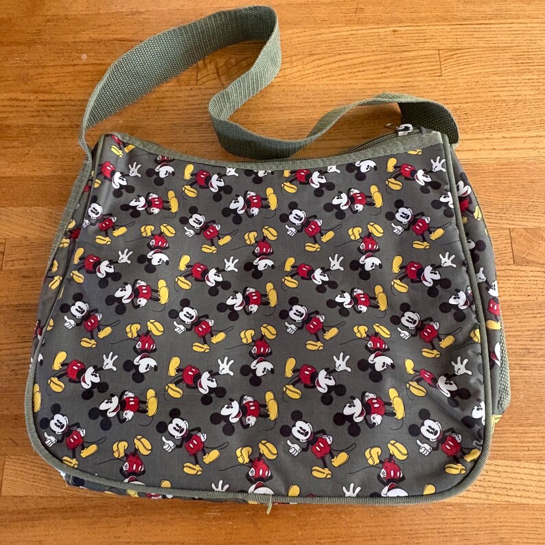 Puede incluir: Un bolso bandolera gris con un patr&oacute;n repetitivo de personajes de Mickey Mouse en rojo, negro, blanco y amarillo. El bolso incluye una correa de hombro ajustable a juego en color verde oliva y un cierre de cremallera en la parte superior.