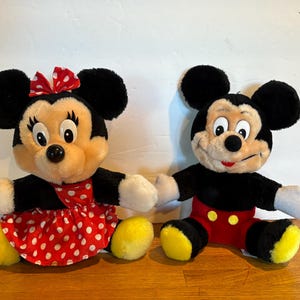 May include: Two plush Disney character dolls, Mickey Mouse and Minnie Mouse, are seated side-by-side. Minnie wears a red polka-dot dress and bow. Mickey wears red shorts and yellow shoes. Both have black ears and are holding hands.