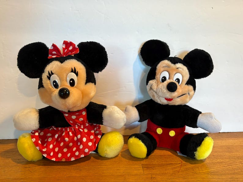 May include: Two plush Disney character dolls, Mickey Mouse and Minnie Mouse, are seated side-by-side. Minnie wears a red polka-dot dress and bow. Mickey wears red shorts and yellow shoes. Both have black ears and are holding hands.