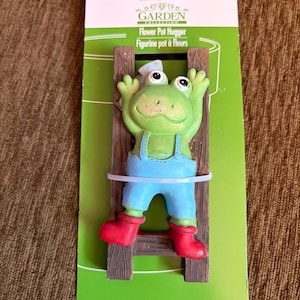 May include: A green frog figurine wearing blue overalls and red boots, clinging to a brown wooden ladder. The figurine is a flower pot hugger, with the words "Flower Pot Hugger" printed on the packaging.