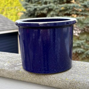 Puede incluir: Una maceta cilíndrica de cerámica azul cobalto con borde blanco. La maceta tiene un acabado brillante y está sobre una superficie de hormigón. El fondo presenta follaje verde y un vistazo a un edificio.