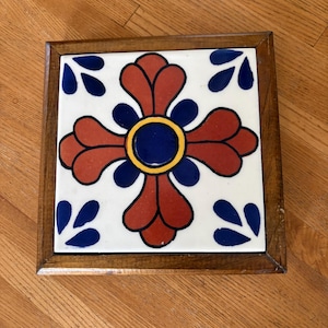 Può includere: Una piastrella in ceramica quadrata inserita in una cornice di legno marrone scuro. La piastrella presenta uno sfondo bianco con un motivo floreale centrale in rosso scuro, blu scuro e giallo, accentuato da quattro motivi a foglia blu negli angoli.