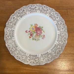 Puede incluir: Un plato redondo blanco con borde festoneado y diseño floral. El plato presenta un borde de filigrana dorada y un ramo central de flores rosas, amarillas y rojas. Una pieza vintage para servir.